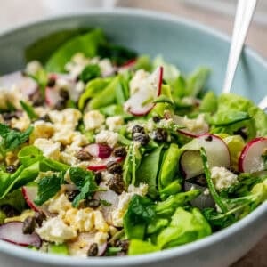 A bowl of green salad topped with sliced radishes, crumbled cheese, and capers, with a spoon resting in the bowl.
