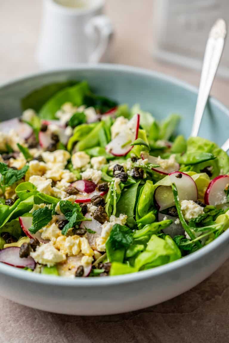A bowl of green salad topped with sliced radishes, crumbled cheese, and capers, with a spoon resting in the bowl.