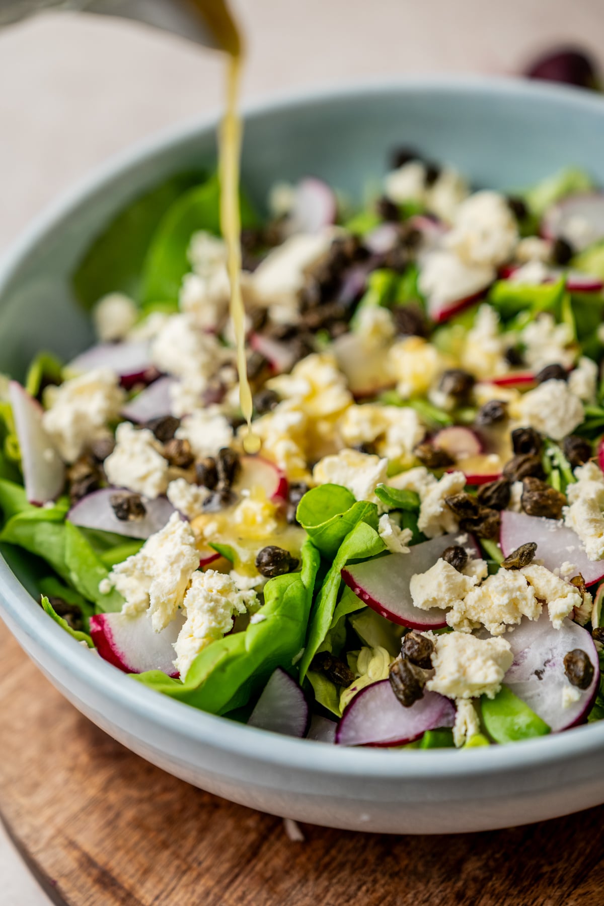 Dressing being poured over a salad of greens, sliced radishes, crumbled feta cheese, and capers in a light blue bowl.