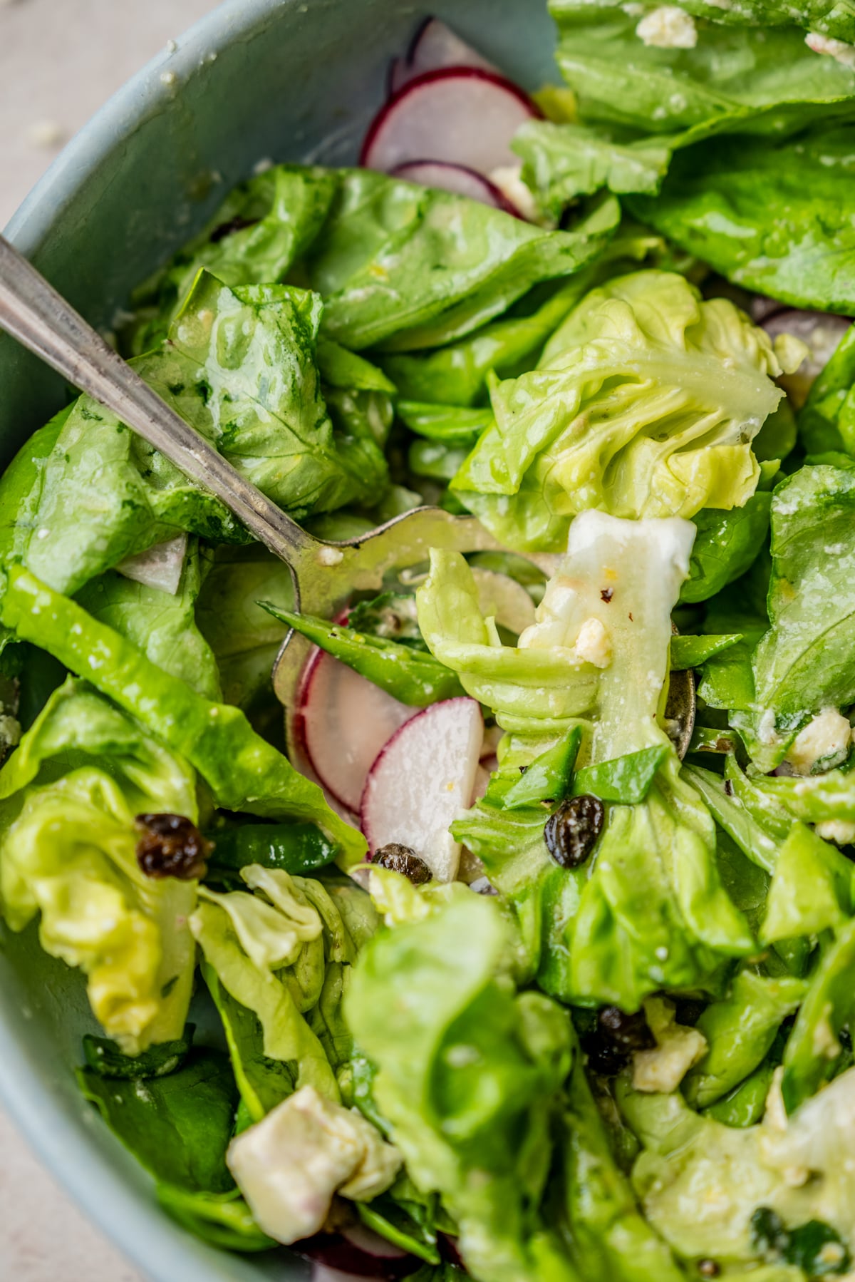 A bowl of green salad with sliced radishes, raisins, and crumbled cheese, being tossed with a metal spoon.