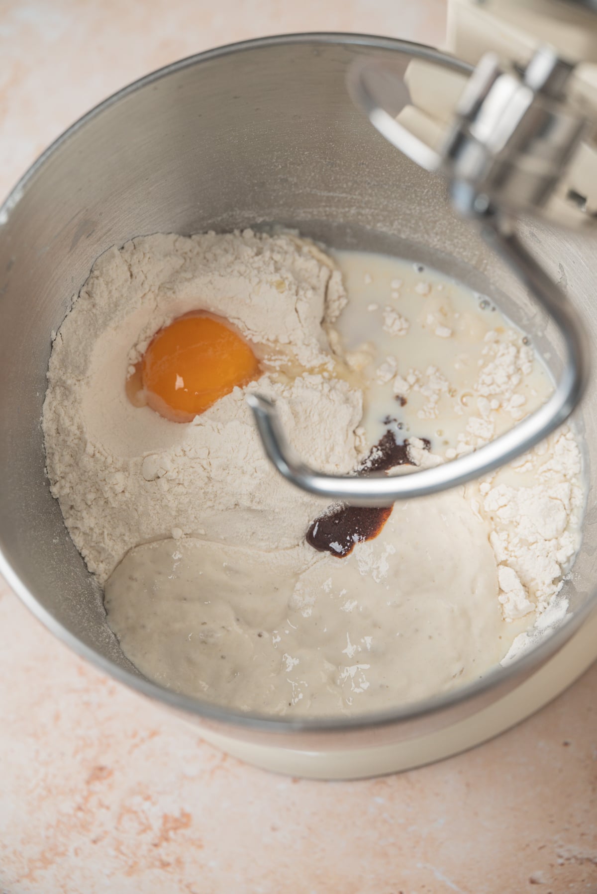 A stand mixer bowl containing flour, a raw egg, milk, and a dollop of sourdough starter with a dough hook attached.